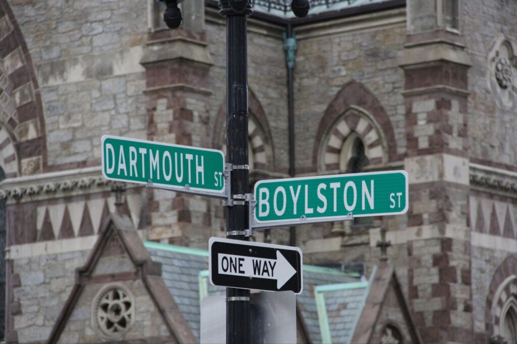 Street corner showing green Dartmouth St and Boylston St signs with one way arrow sign below, historic stone church with arched windows in background